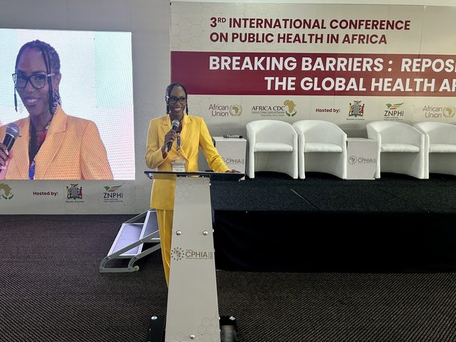 Olivia Otieno in a yellow suit speaking at a podium at the 3rd International Conference on Public Health in Africa
